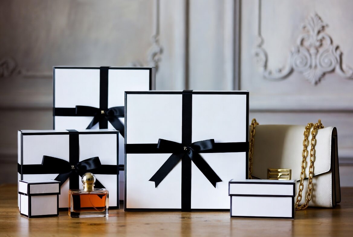 Elegant arrangement of white gift boxes with black satin ribbons and bows on a wooden table, featuring a perfume bottle with amber liquid and a white handbag with gold chain strap, set against an ornate classical wall.