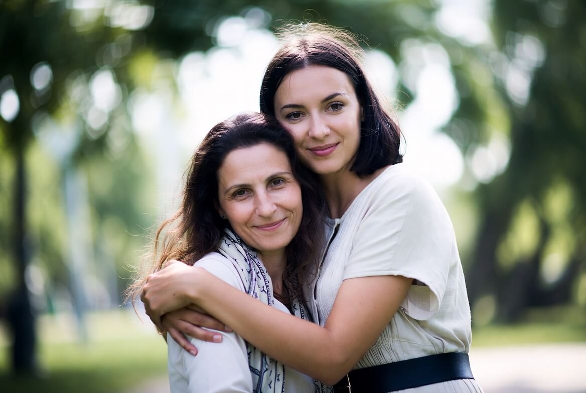 A younger woman gently hugging an older woman from behind; both are smiling warmly with eyes closed in a cozy, loving moment.
